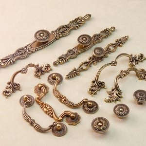 Peut inclure: Un assortiment de quincaillerie de meubles en laiton antique, comprenant des poignées et des boutons ornés. La quincaillerie présente des motifs floraux et des volutes, avec une finition chaude et vieillie. Différentes tailles et styles sont présentés, en centimètres.