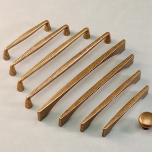 Peut inclure: Une collection de quincaillerie de meubles en laiton antique, comprenant plusieurs poignées et un bouton. Les poignées varient en taille et en design, avec une finition chaude et vieillie. La quincaillerie est disposée sur une surface gris clair.