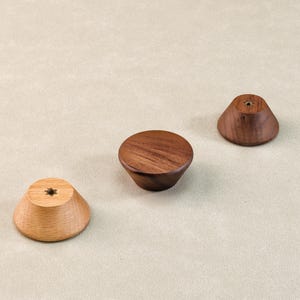 Może przedstawiać: Trzy drewniane gałki do szafek w r&oacute;żnych odcieniach brązu. Gałki mają kształt stożkowy z płaskim wierzchem i otworem do montażu. Usłojenie drewna jest widoczne na każdej gałce, co dodaje im naturalnej estetyki. Gałki te nadają się do mebli.