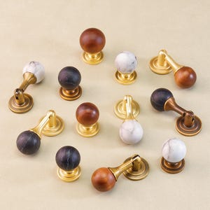 Könnte beinhalten: Eine Sammlung dekorativer Schrankknöpfe und -griffe in verschiedenen Stilen. Die Knöpfe weisen runde, marmorne und hölzerne Designs mit Messing- oder goldfarbener Hardware auf. Die Anordnung zeigt verschiedene Texturen und Farben, passend für Möbel oder Heimdekoration.