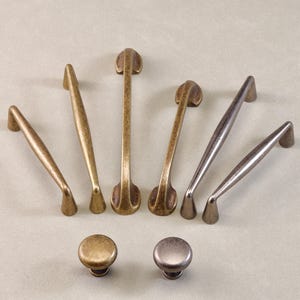 Op de afbeelding: Een verzameling kastbeslag, waaronder verschillende bronzen en zilverkleurige handgrepen en twee bijpassende knoppen. De handgrepen variëren in vorm en grootte, met een mix van gebogen en rechte ontwerpen. Het beslag is geplaatst op een neutrale achtergrond.