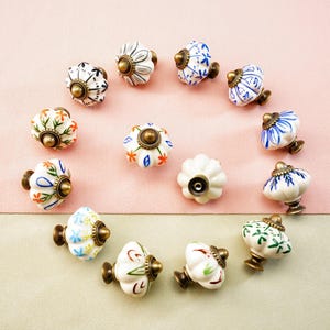 May include: A collection of decorative ceramic cabinet knobs arranged in a circle. The knobs feature various floral and geometric patterns in white, blue, orange, and green, with brass-colored hardware. The background is a two-tone pink and beige.