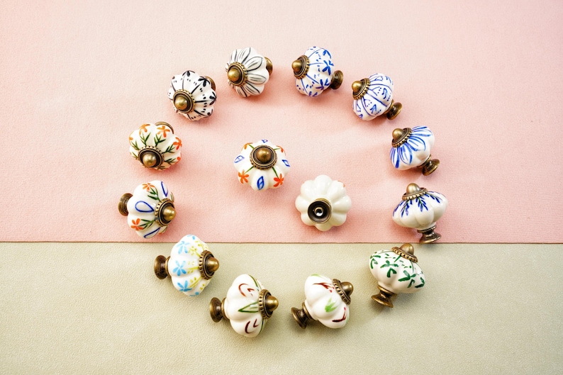 May include: A selection of decorative ceramic cabinet knobs arranged in a circular pattern. The knobs display floral and geometric designs in white, blue, orange, and green, with brass-coloured fittings. The background is a two-tone pink and beige.