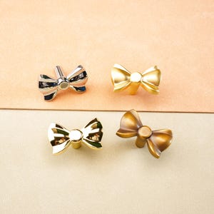 May include: Four bow-shaped cabinet knobs in silver, gold, and bronze finishes. The knobs are arranged on a neutral background, showcasing their decorative design. These knobs are suitable for adding a touch of elegance to furniture.