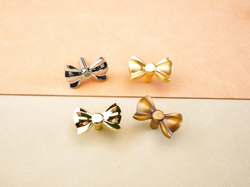 May include: Four bow-shaped cabinet knobs in silver, gold, and bronze finishes. The knobs are arranged on a neutral background, showcasing their decorative design. These knobs are suitable for adding a touch of elegance to furniture.