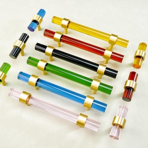 May include: A collection of colorful cabinet pulls in various shades including yellow, red, black, green, blue, and pink. Each pull features a cylindrical acrylic handle with gold-toned metal accents. The pulls are arranged on a light-colored surface.