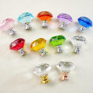 Peut inclure: Une collection de boutons de meuble en cristal coloré et facetté. Les boutons sont de différentes couleurs, dont rose, bleu, orange, violet, rouge, jaune, vert et transparent. Chaque bouton a une base argentée ou dorée.