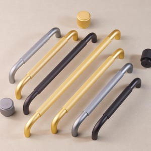 May include: A set of seven cabinet door pulls in various finishes, including gold, silver, and black. The pulls have a textured, ribbed design and a curved shape.