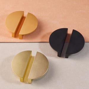 May include: Three cabinet pulls in different finishes. The pulls are made of two half-circle pieces that fit together. The pulls are shown on a light brown surface, a light gray surface, and a dark gray surface. The pulls are gold, brass, and black.