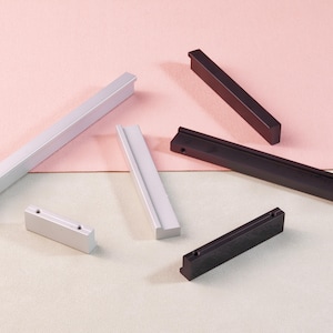 May include: An assortment of modern cabinet pulls in silver and black. The pulls are rectangular and vary in size, with a clean, minimalist design. The background is a soft pink and white.