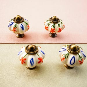 May include: Four decorative cabinet knobs. Each knob features a white ceramic pumpkin shape with red and blue floral accents, a brass-coloured base, and a brass-coloured top. The knobs are arranged on a pink and beige surface.
