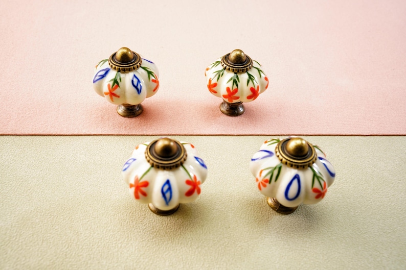 May include: Four decorative cabinet knobs. Each knob features a white ceramic pumpkin shape with red and blue floral accents, a brass-coloured base, and a brass-coloured top. The knobs are arranged on a pink and beige surface.