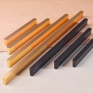 May include: A set of gold and black cabinet pulls. The pulls are rectangular and have a slightly rounded edge. The gold pulls are arranged in a staggered pattern, while the black pulls are arranged in a straight line.
