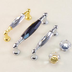 May include: A collection of decorative cabinet hardware, including crystal-like handles and knobs. The handles feature gold and silver-toned bases with faceted crystal centers. Two round knobs are also visible, one with a gold base and one with a silver base.