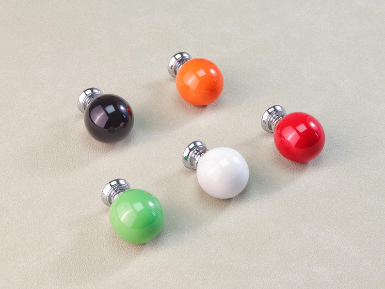 Peut inclure: Cinq boutons de meuble color&eacute;s avec un design rond et brillant. Les boutons sont de diff&eacute;rentes couleurs : noir, orange, rouge, blanc et vert. Chaque bouton a une base argent&eacute;e.