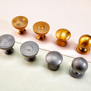 Könnte beinhalten: Eine Auswahl an dekorativen Schrankknöpfen in verschiedenen Metallausführungen. Die Knöpfe umfassen Gold- und Silbertöne mit Designs, die von strukturierten Mustern bis zu glatten, abgerundeten Formen reichen. Diese Hardware-Teile sind für Möbel und Heimdekoration gedacht.