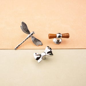 May include: A selection of decorative cabinet hardware. Features a silver dragonfly, a silver bow-shaped knob, and a knob with a silver base and wooden handle. The items are displayed on a two-tone beige and tan background.