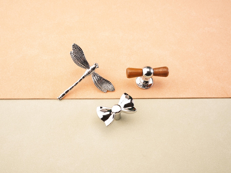 May include: A selection of decorative cabinet hardware. Features a silver dragonfly, a silver bow-shaped knob, and a knob with a silver base and wooden handle. The items are displayed on a two-tone beige and tan background.
