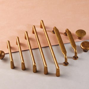 May include: A collection of antique brass cabinet hardware, including knobs and various sizes of pull handles. The hardware is arranged on a two-tone background of pink and gray, showcasing the different designs and sizes available.