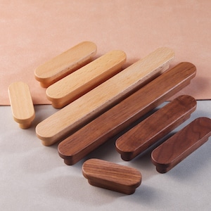 May include: A set of eight wooden cabinet pulls in two different wood tones. The pulls are oval shaped and have a smooth finish. The pulls are arranged in a staggered pattern on a light gray surface.