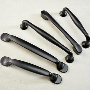 Könnte beinhalten: Eine Sammlung von mattschwarzen Schrankgriffen in verschiedenen gebogenen und geraden Designs. Die Hardware ist auf einer hellen Oberfläche angeordnet, die die verschiedenen Stile und Formen zeigt. Diese Griffe sind ideal für Küchen- oder Badezimmerschränke.