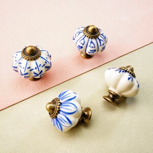 May include: Four decorative ceramic cabinet knobs. Each knob has a white base with blue floral patterns, a brass-coloured metal base, and a brass-coloured top. The knobs are arranged on a pink and green surface.