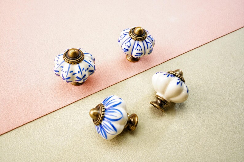 May include: Four decorative ceramic cabinet knobs. Each knob has a white base with blue floral patterns, a brass-coloured metal base, and a brass-coloured top. The knobs are arranged on a pink and green surface.