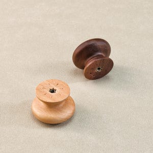 Może przedstawiać: Dwa drewniane gałki do szafek. Jedna jest w kolorze jasnobrązowym, druga ciemnobrązowym. Obie mają zaokrągloną g&oacute;rę i płaską podstawę z otworem montażowym. Gałki są wykonane z drewna i mają prosty, klasyczny design.