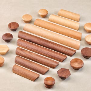May include: An assortment of wooden cabinet knobs and pulls in various shapes and wood tones. The collection includes round, hexagonal, and rectangular designs, showcasing a range of light to dark brown hues. The knobs and pulls are arranged on a neutral surface.