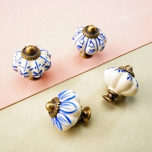 May include: Four decorative ceramic cabinet knobs. Each knob has a white base with blue floral patterns, a brass-coloured metal base, and a brass-coloured top. The knobs are arranged on a pink and green surface.