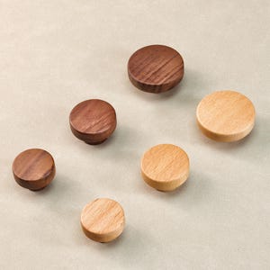 Może przedstawiać: Sześć drewnianych gałek do szafek w r&oacute;żnych odcieniach brązu i naturalnego drewna. Gałki są okrągłe, z płaskim wierzchem i lekko zaokrągloną podstawą. Usłojenie drewna jest widoczne na ciemniejszych gałkach. Wymiary w centymetrach.