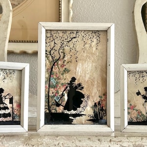 May include: Three framed silhouette art pieces. The frames are white, and the art features black silhouettes of figures in a garden setting. The backgrounds have floral and tree designs. The art pieces are displayed on a white surface.