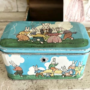 May include: A vintage rectangular tin box with a light blue background and illustrations of rabbits. The lid and sides feature scenes of rabbits in various activities, with the words "PETER RABBIT" on the lid.