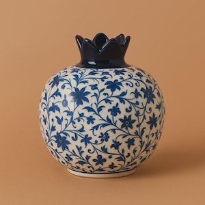 Può includere: Un vaso in ceramica a forma di melograno, con base color crema e corona blu scuro. Il vaso è decorato con un motivo floreale blu. Il design presenta un motivo dettagliato e ripetitivo di fiori e tralci, creando un elegante elemento decorativo.