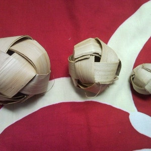 May include: Three woven palm leaf spheres of varying sizes rest on a red and white patterned surface. The spheres are constructed from interwoven strips of light brown palm leaves, creating a textured, open design. The background is a vibrant red with white floral shapes.