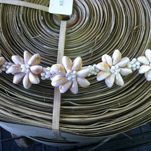 May include: A close-up of a white and gold shell flower lei. The lei is made of small, white shells that are arranged in a flower shape. The shells are accented with gold paint.