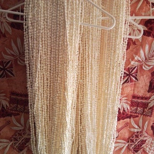 May include: Two white shell leis hanging on a white hanger. The leis are made of small, round, white shells. The leis are hanging against a red and white patterned background.