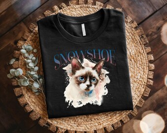 May include: Black t-shirt with a watercolor illustration of a Siamese cat with blue eyes. The cat is surrounded by a white splatter paint effect. The text "SNOWSHOE" is printed in pink and blue above the cat.
