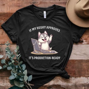 Puede incluir: Camiseta negra con un husky de dibujos animados sentado frente a un ordenador. El texto de la camiseta dice "Si mi husky lo aprueba, está listo para la producción."