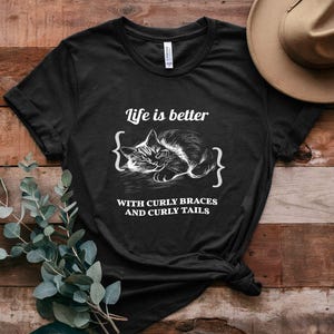 Puede incluir: Una camiseta negra con un gráfico blanco de un gato durmiendo con el texto "Life is better { } with curly braces and curly tails".