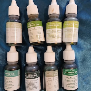 May include: Eight small glass bottles of Stampin' Ink in various shades of green and other colors. Each bottle has a white dropper top and a label with the ink color name. The bottles are arranged on a blue surface.