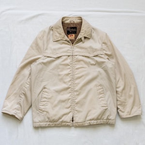 May include: A beige zip-up jacket with a collared neckline and two front pockets. The jacket has a light brown, fuzzy lining around the collar. The brand name "Ashbrook Sportswear" is visible on a tag inside the collar.