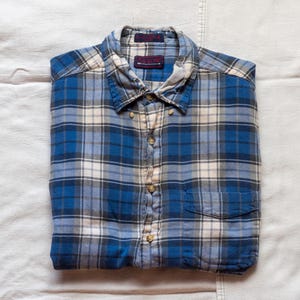 Op de afbeelding: Een blauw, wit en grijs geruit button-down overhemd, netjes opgevouwen. Het overhemd heeft een borstzak en een kraag. De merknaam "ARROW" is zichtbaar op een label bij de kraag. Het overhemd is gemaakt van een zacht, waarschijnlijk flanellen materiaal.