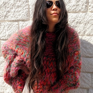 K&ouml;nnte beinhalten: Ein grob gestrickter, &uuml;bergro&szlig;er Pullover mit einer lebendigen, mehrfarbigen Garnmischung aus leuchtendem Pink, Orange und Gr&uuml;n. Der Pullover hat ein dickes, strukturiertes Gewebe mit langen &Auml;rmeln und einem Rundhalsausschnitt &uuml;ber einem hellen Kleidungsst&uuml;ck.