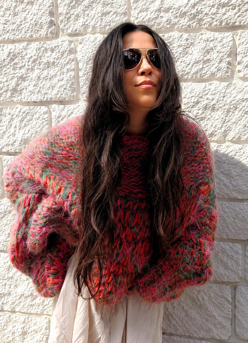K&ouml;nnte beinhalten: Ein grob gestrickter, &uuml;bergro&szlig;er Pullover mit einer lebendigen, mehrfarbigen Garnmischung aus leuchtendem Pink, Orange und Gr&uuml;n. Der Pullover hat ein dickes, strukturiertes Gewebe mit langen &Auml;rmeln und einem Rundhalsausschnitt &uuml;ber einem hellen Kleidungsst&uuml;ck.