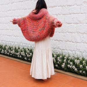 K&ouml;nnte beinhalten: Eine &uuml;bergro&szlig;e, flauschige, handgestrickte Strickjacke in leuchtenden Rosa-, Orange- und Gr&uuml;nt&ouml;nen mit einer lockeren, volumin&ouml;sen Silhouette und langen &Auml;rmeln, kombiniert mit einem bodenlangen, einfarbigen, cremefarbenen Stufenrock.