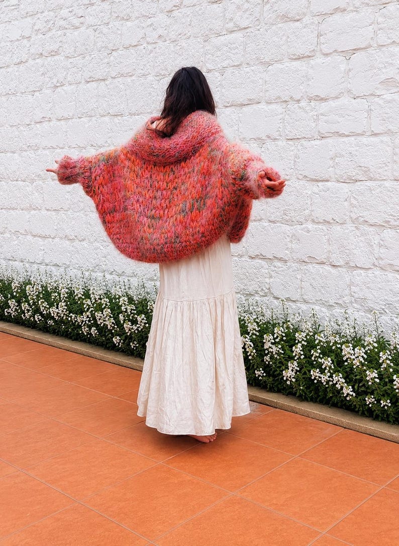 K&ouml;nnte beinhalten: Eine &uuml;bergro&szlig;e, flauschige, handgestrickte Strickjacke in leuchtenden Rosa-, Orange- und Gr&uuml;nt&ouml;nen mit einer lockeren, volumin&ouml;sen Silhouette und langen &Auml;rmeln, kombiniert mit einem bodenlangen, einfarbigen, cremefarbenen Stufenrock.