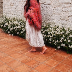 K&ouml;nnte beinhalten: Eine lang&auml;rmelige, flauschige, mehrfarbige Strickjacke in Rosa-, Orange- und Rott&ouml;nen, getragen &uuml;ber einem schlichten, hellen, wadenlangen Rock. Die Strickjacke zeichnet sich durch eine dicke, strukturierte Strickkonstruktion mit lockerer Passform aus.
