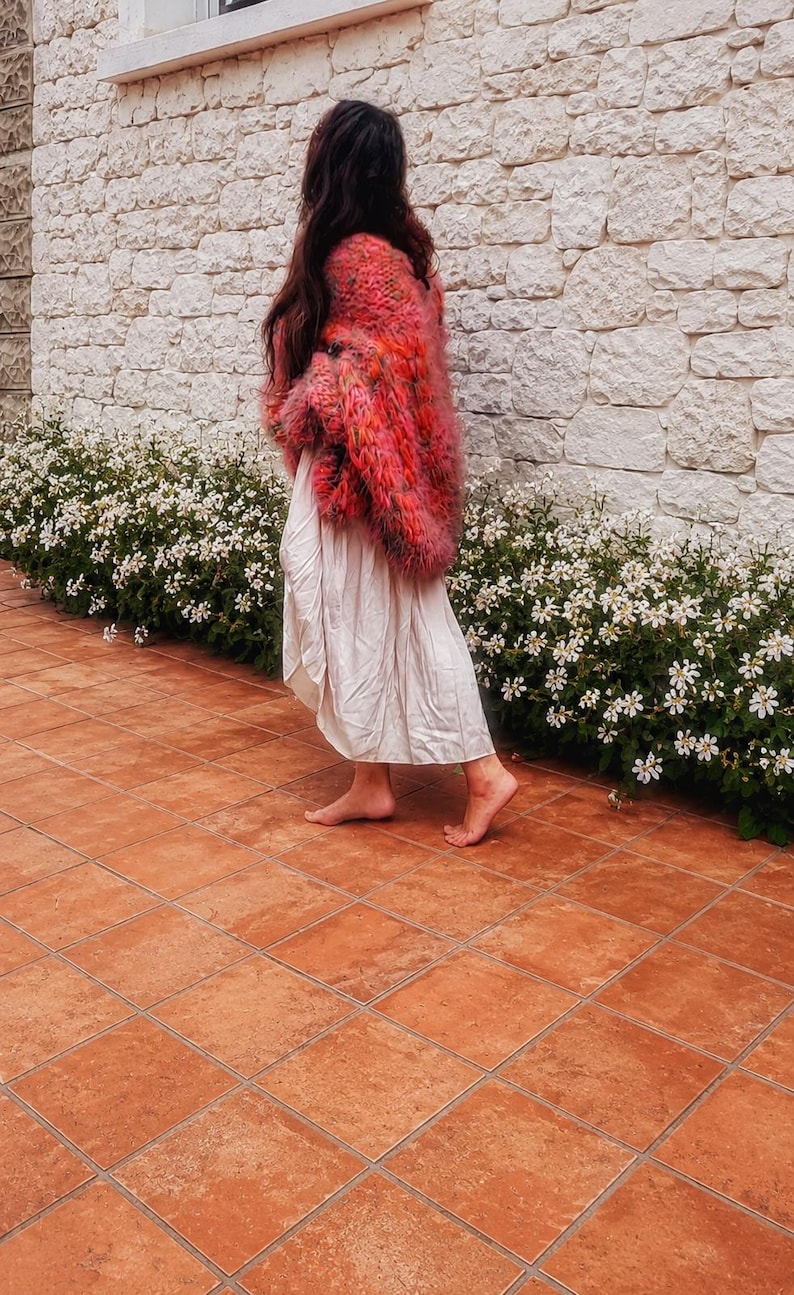 K&ouml;nnte beinhalten: Eine lang&auml;rmelige, flauschige, mehrfarbige Strickjacke in Rosa-, Orange- und Rott&ouml;nen, getragen &uuml;ber einem schlichten, hellen, wadenlangen Rock. Die Strickjacke zeichnet sich durch eine dicke, strukturierte Strickkonstruktion mit lockerer Passform aus.