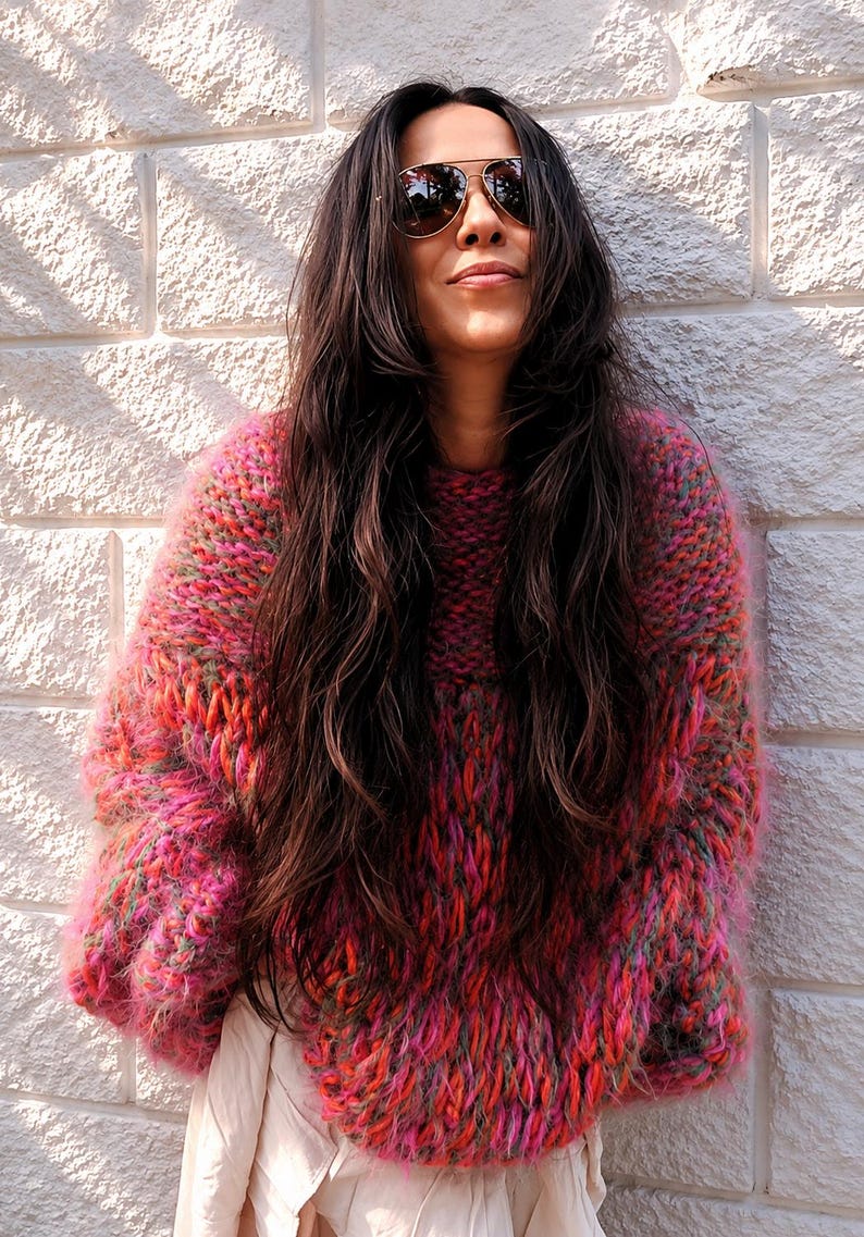 K&ouml;nnte beinhalten: Ein grob gestrickter, &uuml;bergro&szlig;er Pullover mit einer lebendigen, mehrfarbigen Garnmischung aus leuchtendem Pink, Orange und Gr&uuml;n. Das Kleidungsst&uuml;ck hat ein dickes, strukturiertes Gewebe mit flauschiger Optik, langen &Auml;rmeln und einem Rundhalsausschnitt.